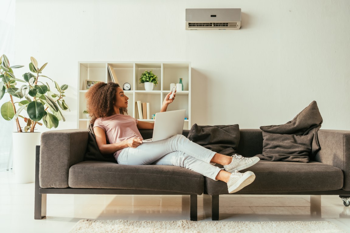 pretty african american woman using laptop and holding remote controller while lying on sofa under